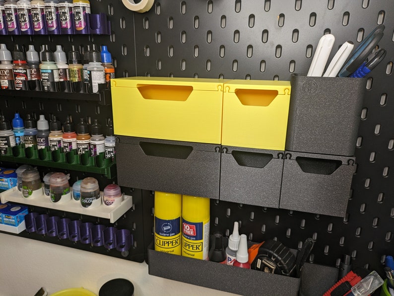 May include: A pegboard wall with various storage solutions, including yellow plastic drawers, black plastic bins, and a shelf with small bottles. The pegboard is black with a grid pattern.