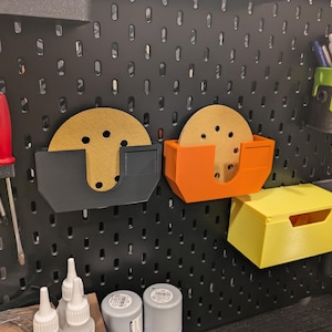 May include: A black pegboard wall with various tools and storage containers. There are two orange and black sanding disc holders, a yellow storage bin, and a black tool holder with a variety of tools.