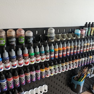 May include: A black pegboard with rows of small paint bottles in various colours. The bottles are organised in rows and columns, with some bottles labelled "Citadel Colour" and others labelled "Game Colour".