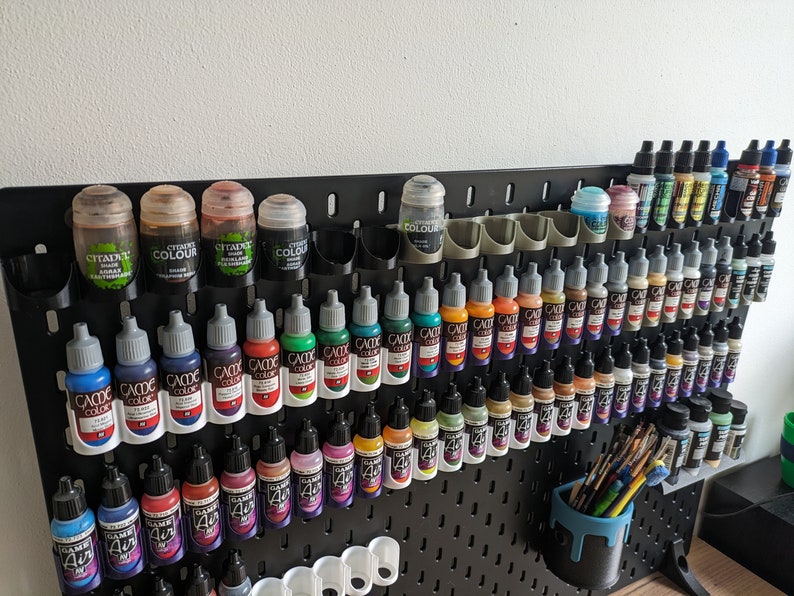 May include: A black pegboard with rows of small paint bottles in various colours. The bottles are organised in rows and columns, with some bottles labelled "Citadel Colour" and others labelled "Game Colour".
