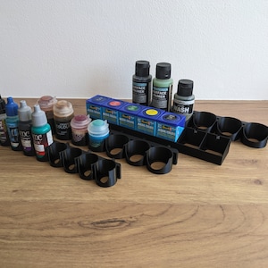 May include: A black plastic paint rack with 10 slots holds various bottles of acrylic paint, including a surface primer, a wash, and several colours. The bottles are arranged in rows and are mostly blue, green, and brown.