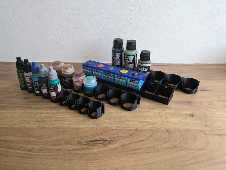 May include: A black plastic paint rack with 10 slots holds various bottles of acrylic paint, including a surface primer, a wash, and several colours. The bottles are arranged in rows and are mostly blue, green, and brown.