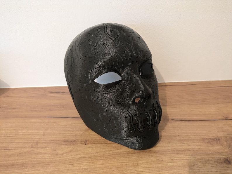 Op de afbeelding: Een zwart schedelmasker met ingewikkelde details en witte ooggaten. Het masker is gemaakt van glad, hard plastic en heeft een getextureerd oppervlak.