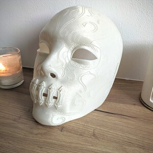 Op de afbeelding: Witte plastic masker met ingewikkelde ontwerpen en een gesloten mond. Het masker ligt op een houten oppervlak met twee brandende kaarsen op de achtergrond.