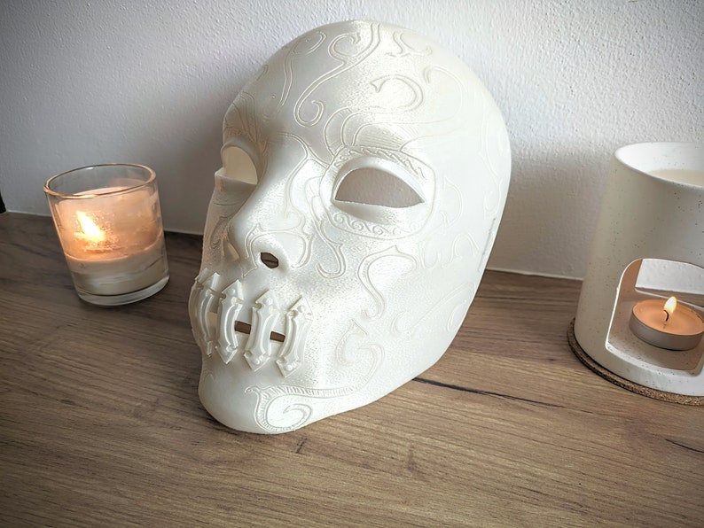 Op de afbeelding: Witte plastic masker met ingewikkelde ontwerpen en een gesloten mond. Het masker ligt op een houten oppervlak met twee brandende kaarsen op de achtergrond.