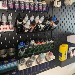 Può includere: Una parete di pannello forato nero con varie bottiglie e contenitori di vernice organizzati su mensole e rack. Le bottiglie sono etichettate con nomi di marchi come "Game Air" e "Vallejo".