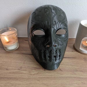 May include: A dark green mask with intricate designs and a skull-like mouth. The mask is made of plastic and has a smooth finish.