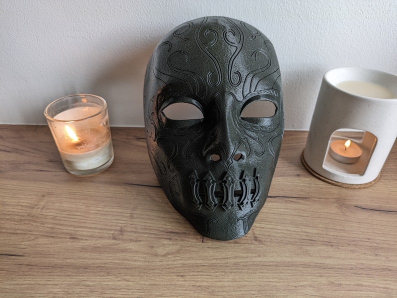 Op de afbeelding: Een donkergroen masker met ingewikkelde ontwerpen en een schedelvormige mond. Het masker is gemaakt van plastic en heeft een gladde afwerking.
