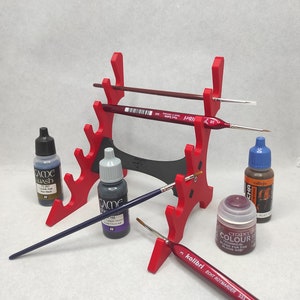 May include: Red plastic paint brush holder with multiple slots for brushes and bottles. The holder has a black base. There are several paint bottles and brushes in the holder. The bottles are labeled "Game Wash", "Game", "Citadel Colour", and "Vallejo".