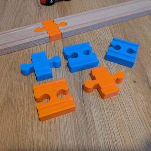 May include: A set of wooden train track pieces including a long straight section and several smaller plastic connectors in bright blue and orange. The connectors feature puzzle-like tabs and slots designed to link the wooden track segments together.