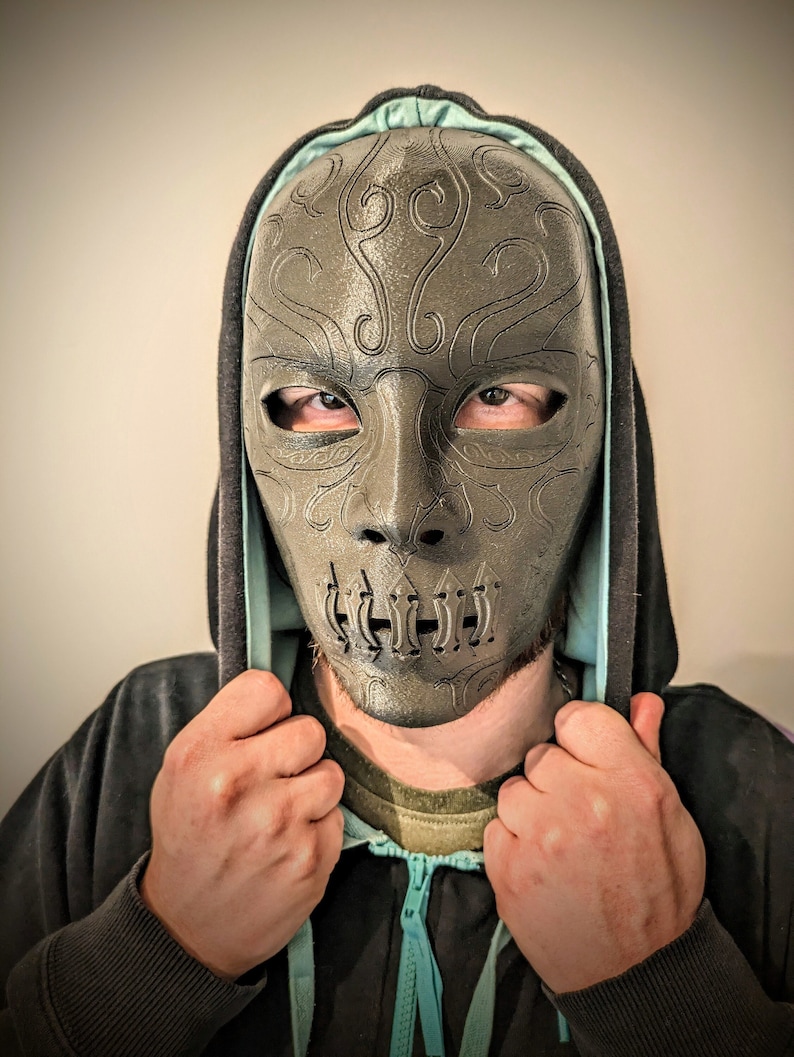 Op de afbeelding: Een persoon draagt een zwarte hoodie met een turquoise rits en een donkergrijs masker met ingewikkelde ontwerpen en een mond met een reeks gaten.