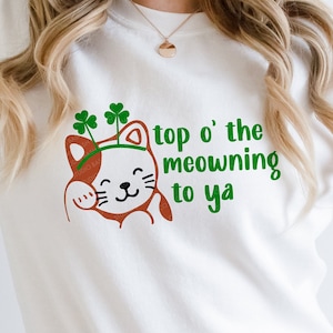 May include: White sweatshirt with a St. Patrick's Day theme. Features a cartoon cat with a green shamrock headband and the phrase "top o' the meowning to ya" in green lettering. A festive and playful design.