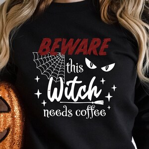 May include: Black sweatshirt with a white graphic that says "Beware this witch needs coffee" with a spiderweb and witch's eyes. The sweatshirt is being held by a person holding a glittery orange pumpkin.