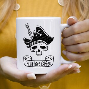 Könnte beinhalten: Weiße Keramik-Kaffeetasse mit einer schwarz-weißen Illustration eines Schädels, der einen Piratenhut trägt und ein Schwert hält. Die Tasse hat eine schwarze Schriftrolle mit dem Text "Must have Coffee" darauf.