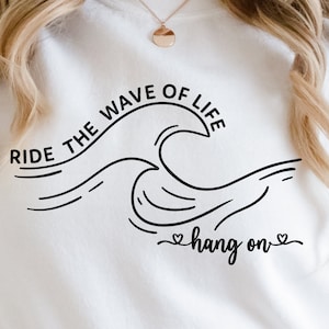 Puede incluir: Sudadera blanca con texto negro que dice "Ride the wave of life hang on" con un gráfico de ola.