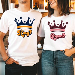 May include: Two white t-shirts with a crown and text. The shirt on the left says "King" in orange and blue lettering. The shirt on the right says "Queen" in pink and blue lettering.
