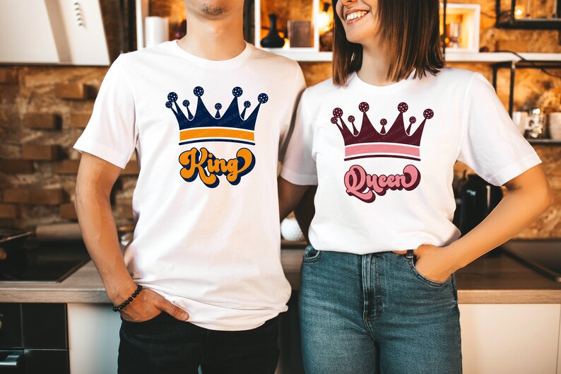 May include: Two white t-shirts with a crown and text. The shirt on the left says "King" in orange and blue lettering. The shirt on the right says "Queen" in pink and blue lettering.