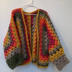 May include: A crocheted cardigan with a colourful striped pattern in shades of green, yellow, orange, and red. The cardigan has a V-neck and long sleeves.