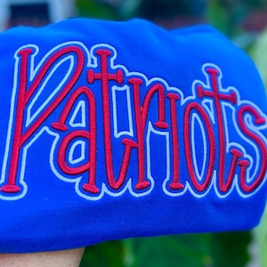 Könnte beinhalten: Königsblaues Sweatshirt mit dem Wort "Patriots" in Rot, weiß umrandet. Der Text ist in einer kursiven, stilisierten Schriftart gehalten. Das Sweatshirt ist aus einem weichen Material.