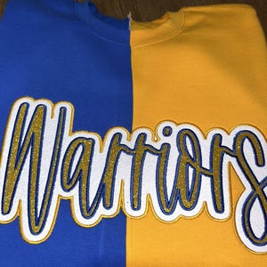 May include: A blue and gold sweatshirt with the word "Warriors" embroidered across the front. The sweatshirt is split vertically, with the left side blue and the right side gold. The word "Warriors" is in a script font with gold glitter.