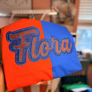 May include: A two-toned t-shirt, split vertically with orange and blue sections. The word "Flora" is embroidered in a glittery orange script, outlined in blue. The shirt is folded, showcasing the design.