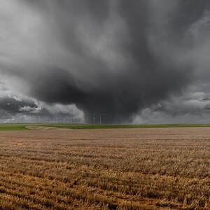 May include: A dramatic landscape photograph featuring a vast, harvested field under a dark, ominous storm cloud. Wind turbines are visible on the horizon, with a strip of green grass separating the field and the sky.