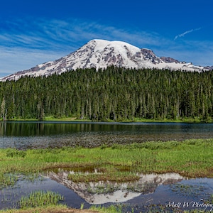 May include: A scenic view of a snow-capped mountain reflected in a still lake. The mountain is surrounded by a dense forest of evergreen trees. The sky is a clear blue with white clouds.