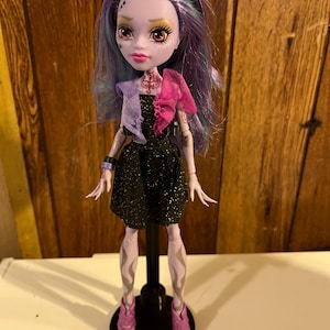 May include: A purple-haired doll with a black dress and pink shoes. The doll has a pale complexion and red eyes. The doll is wearing a black dress with glitter and a pink and purple scarf.