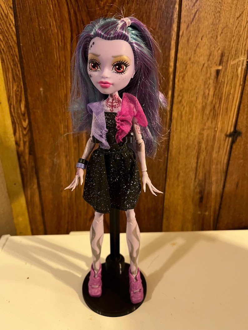 May include: A purple-haired doll with a black dress and pink shoes. The doll has a pale complexion and red eyes. The doll is wearing a black dress with glitter and a pink and purple scarf.