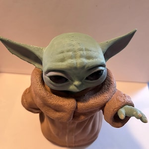 May include: A figurine of a character with large ears and a light green complexion, wearing a brown robe. The figure has large, dark eyes and a small hand extended outward. The figurine is a character from a popular science fiction franchise.