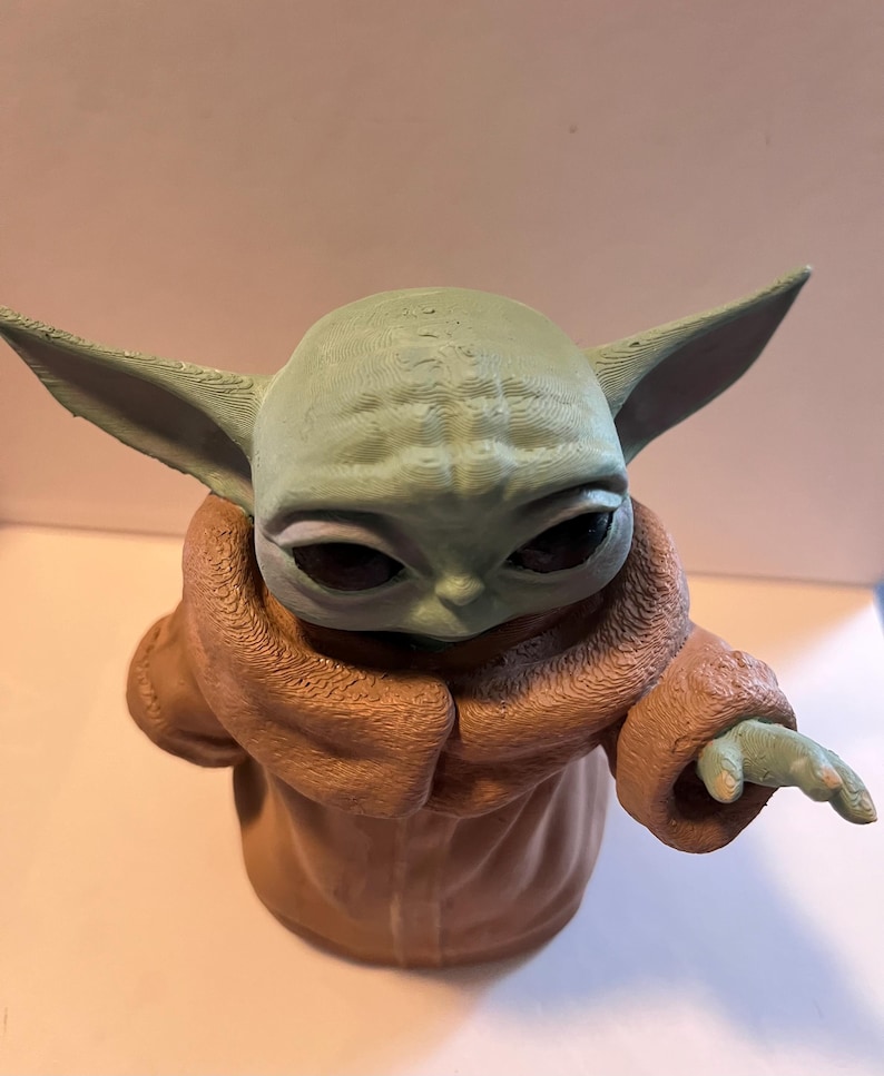 May include: A figurine of a character with large ears and a light green complexion, wearing a brown robe. The figure has large, dark eyes and a small hand extended outward. The figurine is a character from a popular science fiction franchise.
