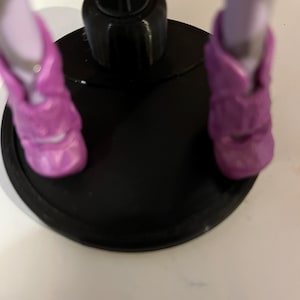 May include: A doll stand with a black base and a black metal rod. The doll is wearing purple shoes.