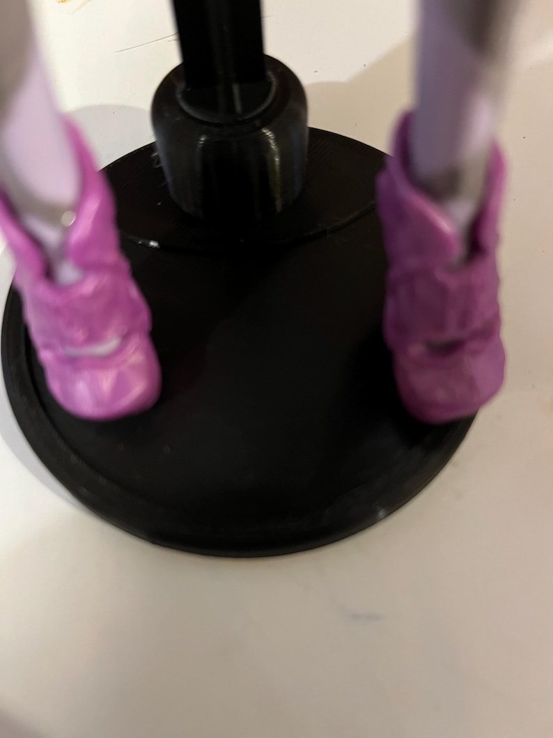 May include: A doll stand with a black base and a black metal rod. The doll is wearing purple shoes.