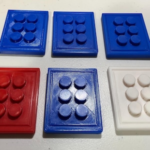 May include: Six rectangular, 3D-printed blocks with raised circular studs. The blocks are in various colors: blue, red, and white. Each block has six studs arranged in two rows of three. The blocks are likely for educational or tactile purposes.