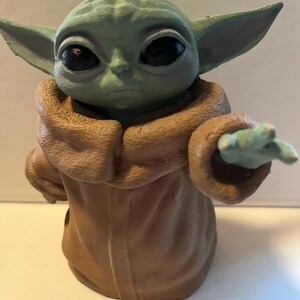 May include: A green and brown figurine of a character with large ears and eyes, wearing a brown robe. The figurine is posed with one hand extended, and the details are clearly visible.