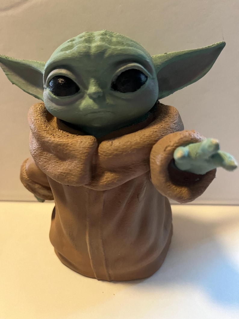 May include: A green and brown figurine of a character with large ears and eyes, wearing a brown robe. The figurine is posed with one hand extended, and the details are clearly visible.