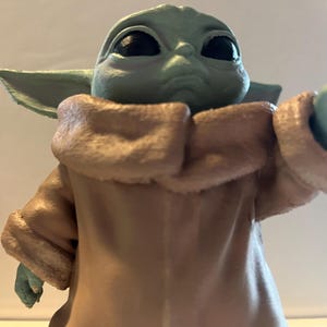 May include: A figurine of a character with large black eyes, green skin, and large ears. The character is wearing a tan coat with a light brown collar. The figurine is a collectible item.