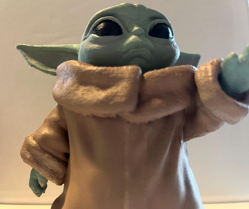 May include: A figurine of a character with large black eyes, green skin, and large ears. The character is wearing a tan coat with a light brown collar. The figurine is a collectible item.