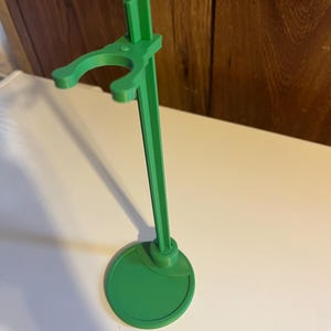 May include: A green plastic doll stand with a round base and a single arm for holding a doll.