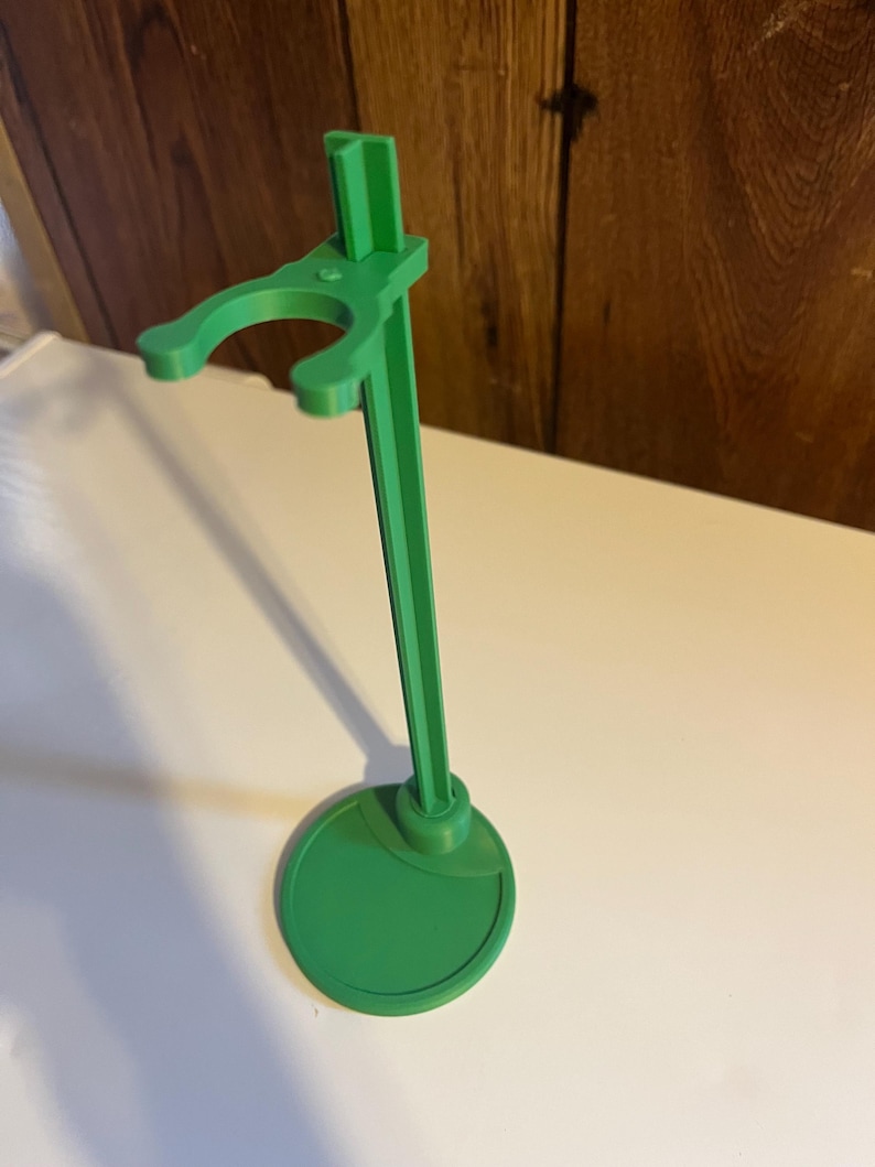 May include: A green plastic doll stand with a round base and a single arm for holding a doll.
