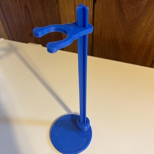 May include: A blue plastic doll stand with a round base and a hook for holding a doll.