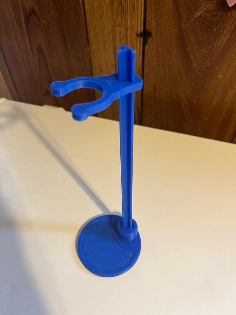 May include: A blue plastic doll stand with a round base and a hook for holding a doll.