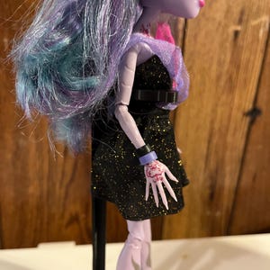 May include: A purple and blue haired doll with a black sparkly dress and a black belt. The doll has a pink and purple shawl and a tattoo on her arm. The doll is standing on a black stand.