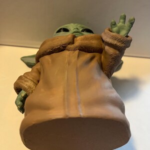 May include: A green figurine of a character from a popular science fiction series. The figure is wearing a tan coat with a brown fur-like collar and cuffs. The figure's hand is raised in a greeting gesture.