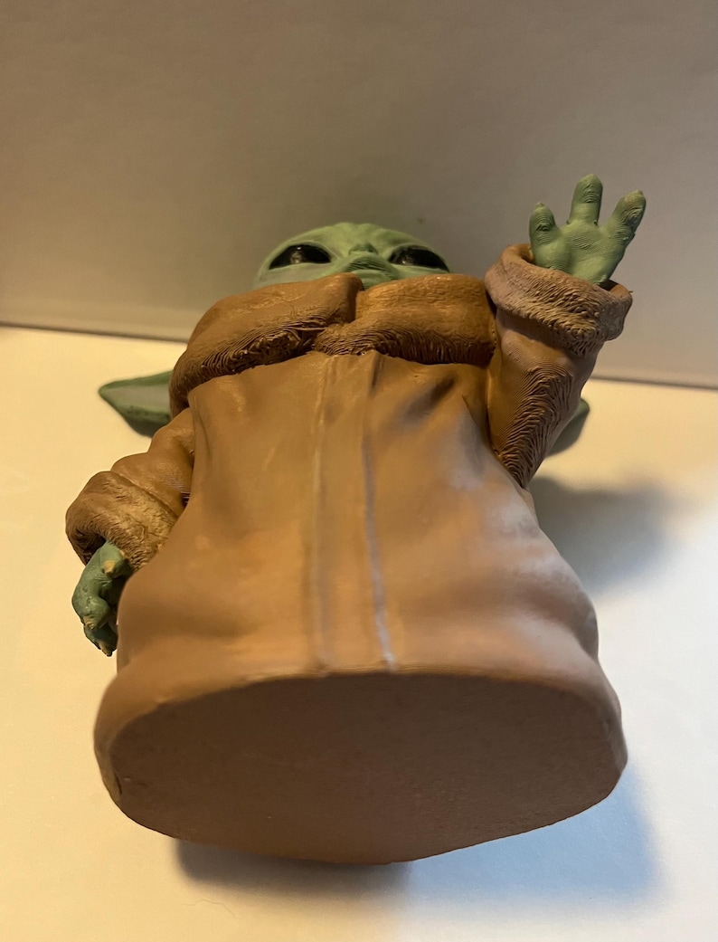 May include: A green figurine of a character from a popular science fiction series. The figure is wearing a tan coat with a brown fur-like collar and cuffs. The figure's hand is raised in a greeting gesture.