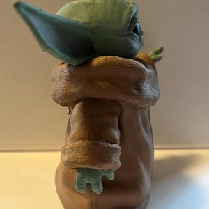 May include: A figurine of a character with large ears, green skin, and a brown coat with a fur-like collar. The figure is posed with one arm raised. The figurine is a collectible item.
