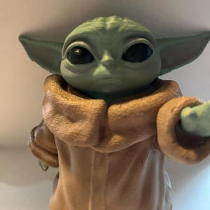 May include: A green figurine of a character with large black eyes and pointed ears. The figure is wearing a tan-colored robe with a hood. The character's skin is a light green color.
