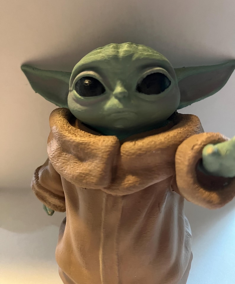 May include: A green figurine of a character with large black eyes and pointed ears. The figure is wearing a tan-colored robe with a hood. The character's skin is a light green color.