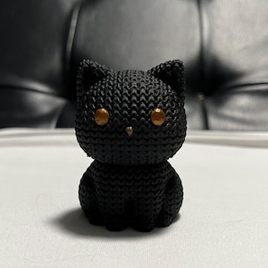 Könnte beinhalten: Eine schwarze Katzenfigur mit goldenen Augen. Die Katze besteht aus einem strukturierten Material, möglicherweise gestrickt oder gehäkelt, und sitzt aufrecht. Die Figur hat ein schlichtes, stilisiertes Design.
