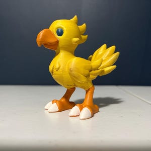 May include: A yellow Chocobo bird figurine with an orange beak, feet, and white-tipped toes. The bird has blue eyes and a spiky crest. The figurine is on a white surface with a dark blue background.